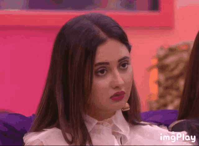a woman in a white shirt is sitting on a couch with a pink wall behind her and the words imgplay visible