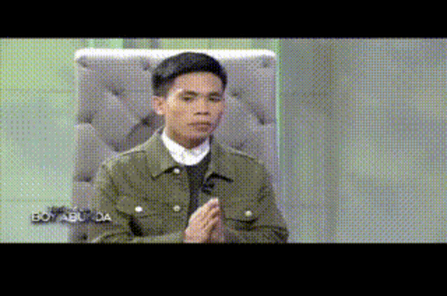 a man in a green jacket is sitting in a chair with his hands folded in front of him .