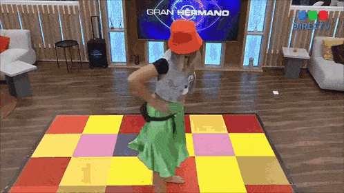 a woman in an orange hat is dancing on a colorful dance floor in front of a tv screen that says gran hermano