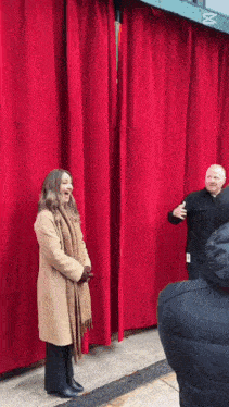 a woman in a tan coat is standing in front of a large red curtain .