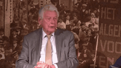 an elderly man in a suit and tie is sitting in front of a crowd of people .