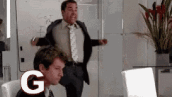 a man in a suit and tie is jumping in the air in front of a white board with the letter g on it .