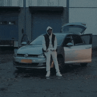 a man wearing a white hoodie and a black vest is standing in front of a car