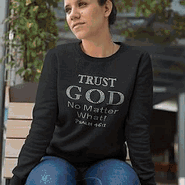 a woman is sitting on a bench wearing a black sweatshirt that says `` trust god no matter what ! ''