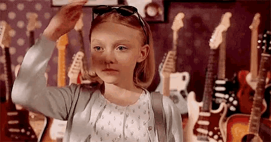 a little girl wearing sunglasses is standing in front of a wall full of guitars .