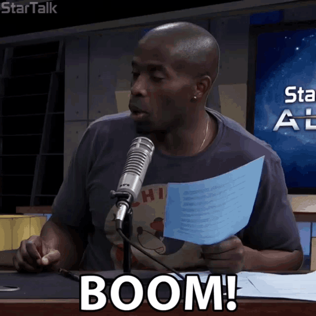 a man speaking into a microphone while holding a piece of paper that says boom on it