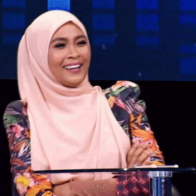 a woman wearing a pink hijab and a floral jacket sits at a table with syiz written on the table