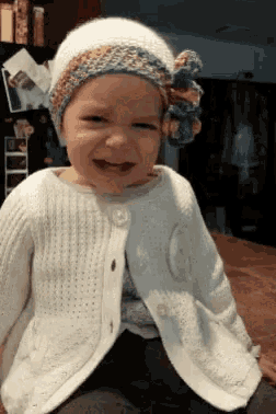 a little girl wearing a white cardigan and a white hat is smiling