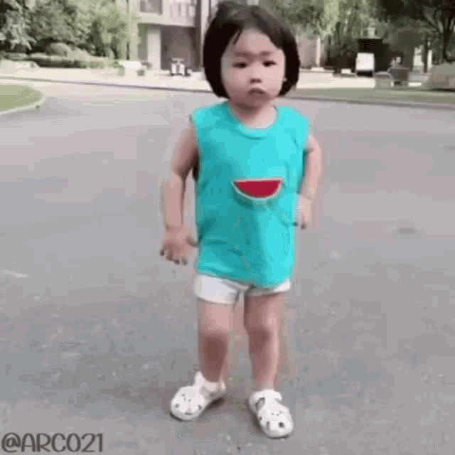 a little girl wearing a blue tank top with a watermelon on it is standing on a street .