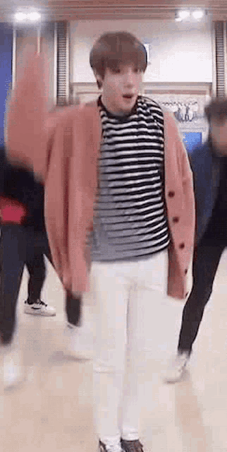 a young man wearing a pink cardigan and a striped shirt is dancing in a room .