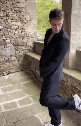 a man in a suit and white sneakers stands on one leg in front of a stone wall