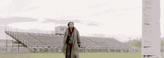 a woman in a trench coat is walking on a field next to a stadium .