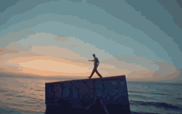 a man is walking on a concrete wall overlooking the ocean at sunset