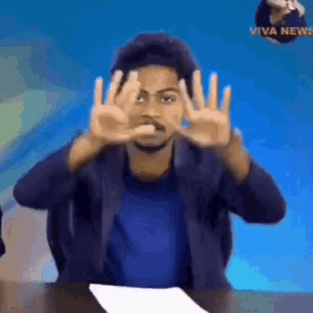 a man is sitting at a table with his hands up in front of a viva news sign .