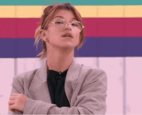 a woman wearing glasses and a striped jacket is standing with her arms crossed in front of a colorful wall .