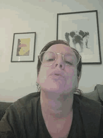 a woman wearing glasses is making a funny face in front of two framed pictures on a wall .