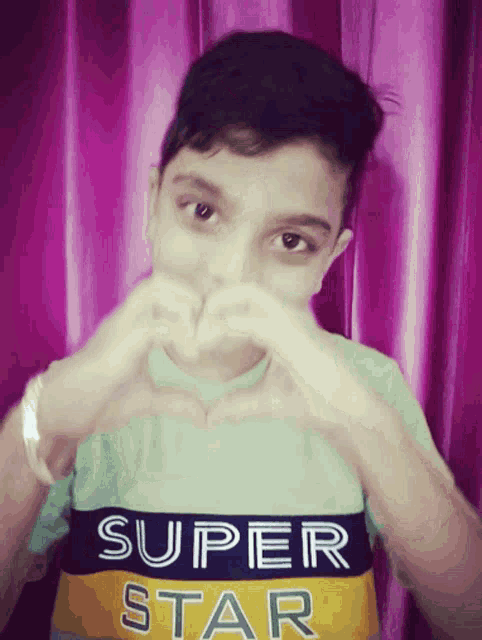 a young boy making a heart shape with his hands wearing a super star shirt