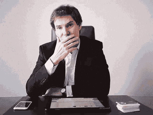 a man in a suit sits at a desk with his hand on his mouth