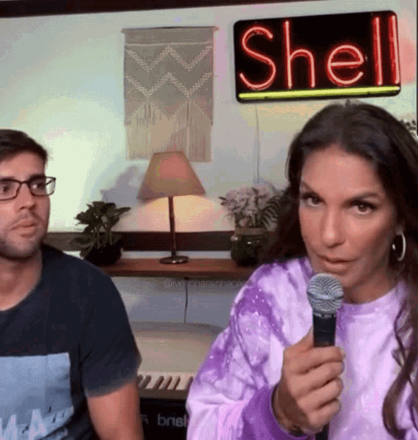 a woman is holding a microphone in front of a shell neon sign