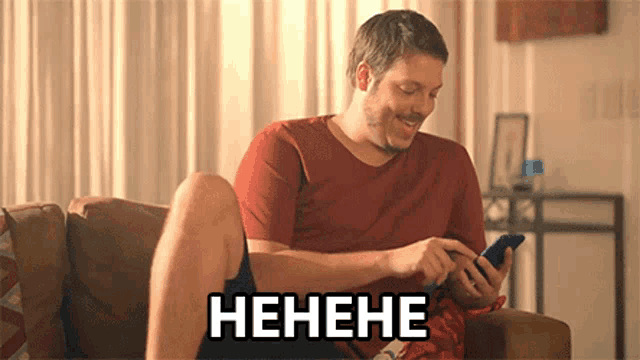 a man is sitting on a couch laughing while looking at his phone with the word henehe above him .