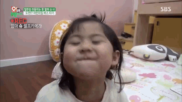 a little girl is making a funny face while sitting on a bed with sbs written on the bottom of the screen