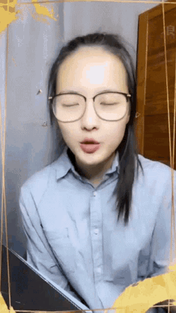 a woman wearing glasses and a blue shirt looks at the camera with her eyes closed