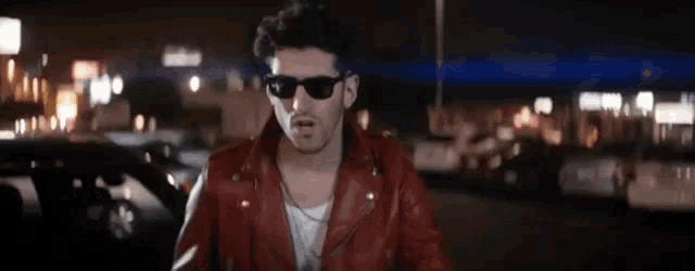 a man in a red leather jacket and sunglasses is standing in a parking lot at night .