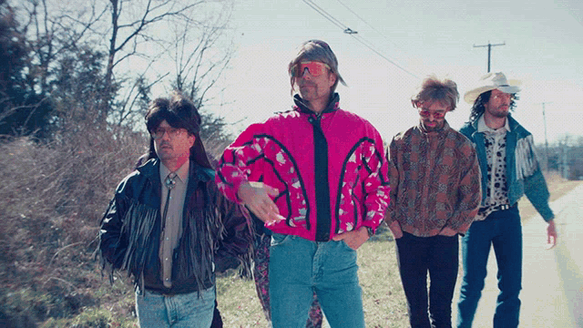 a group of men are walking down a road and one man is wearing a pink jacket
