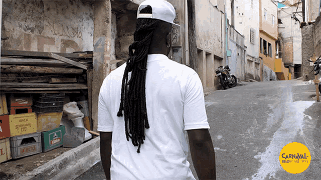 a man in a white shirt is walking down a narrow street with a carnival logo in the corner