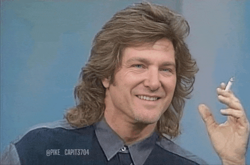 a man with long hair is smiling while holding a cigarette in his hand .