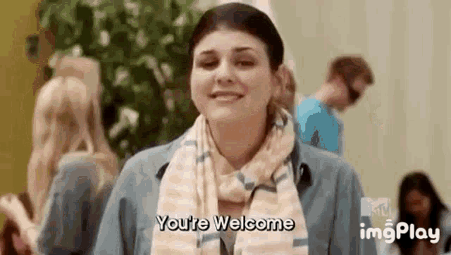 a woman wearing a scarf is smiling and saying `` you 're welcome '' while standing in a crowd of people .
