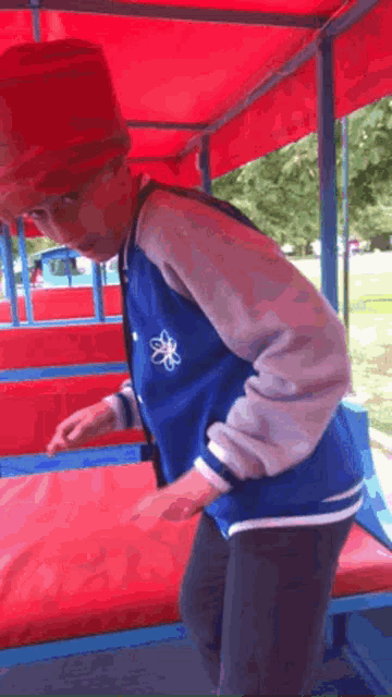 a young boy wearing a blue jacket and a red hat is standing on a red and blue bench .
