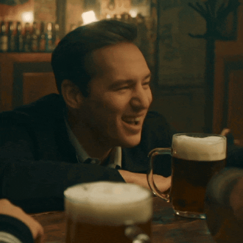a man sits at a table with two mugs of beer and laughs