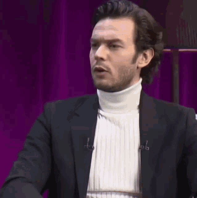 a man wearing a black jacket and a white turtleneck sweater is talking
