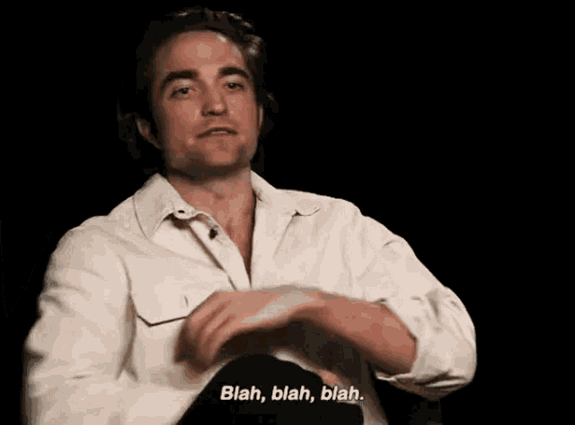 a man in a white shirt is sitting down with his hands folded and talking .