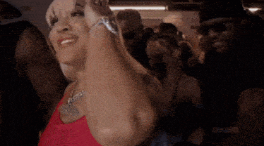 a woman in a red tank top is standing in a crowd of people and giving a peace sign .