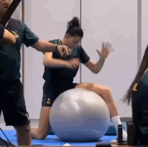 a woman is kneeling on a pilates ball while a man stands behind her .