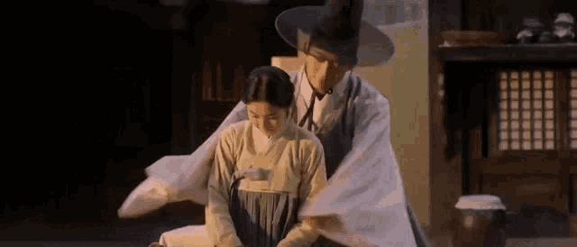 a man in a hat is kneeling down next to a woman in a traditional korean dress .