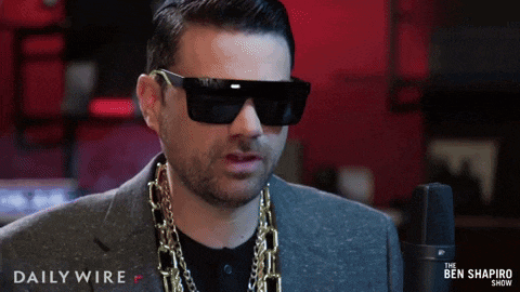a man wearing sunglasses and a gold chain stands in front of a microphone that says daily wire