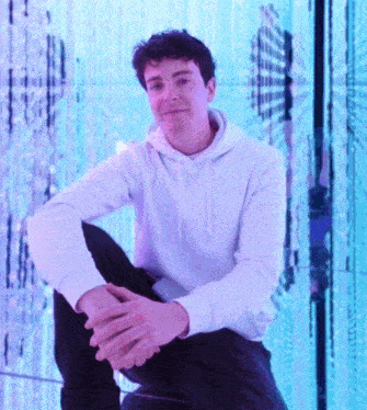 a man in a white hoodie sits with his legs crossed in front of a purple background
