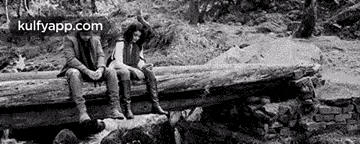 a black and white photo of a man and woman sitting on a log over a river .