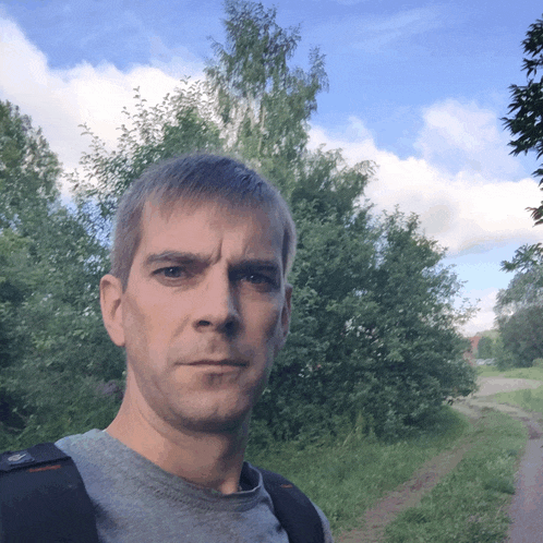 a man wearing a grey shirt and a black backpack stands on a dirt road