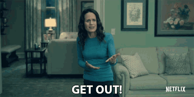a woman in a blue sweater is standing in front of a couch and says get out