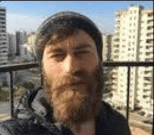 a man with a beard and a hat is standing on a balcony with a city in the background .