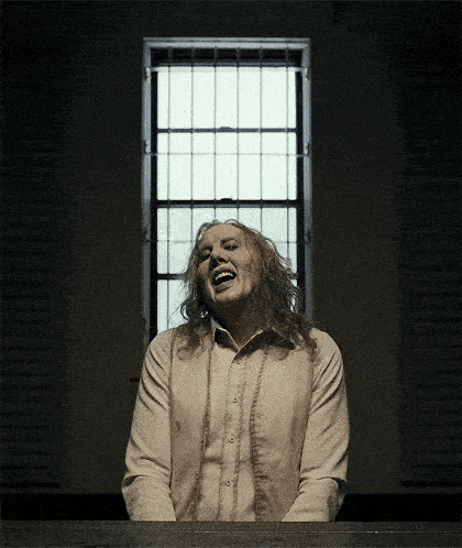 a man with long hair is laughing in front of a window with bars on it