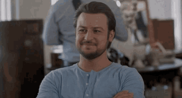 a man with a beard is smiling with his arms crossed in a room