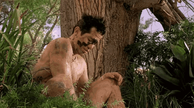 a man with mud on his face sits under a tree in the woods