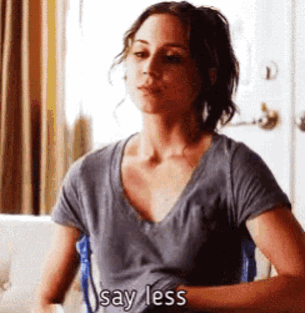 a woman in a grey shirt is sitting on a couch and saying say less