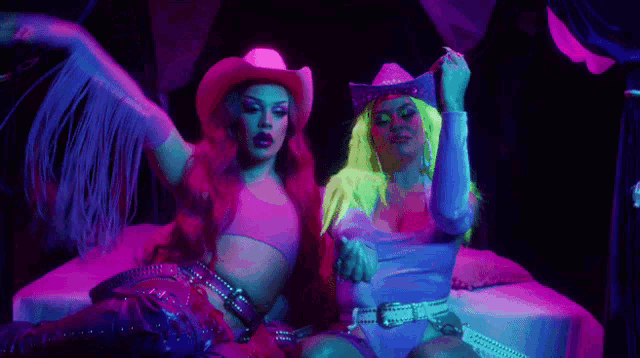 two drag queens in cowboy hats are sitting on a bed in a dark room .