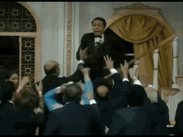 a man in a tuxedo is being lifted by a crowd of people with the word merry on the bottom left